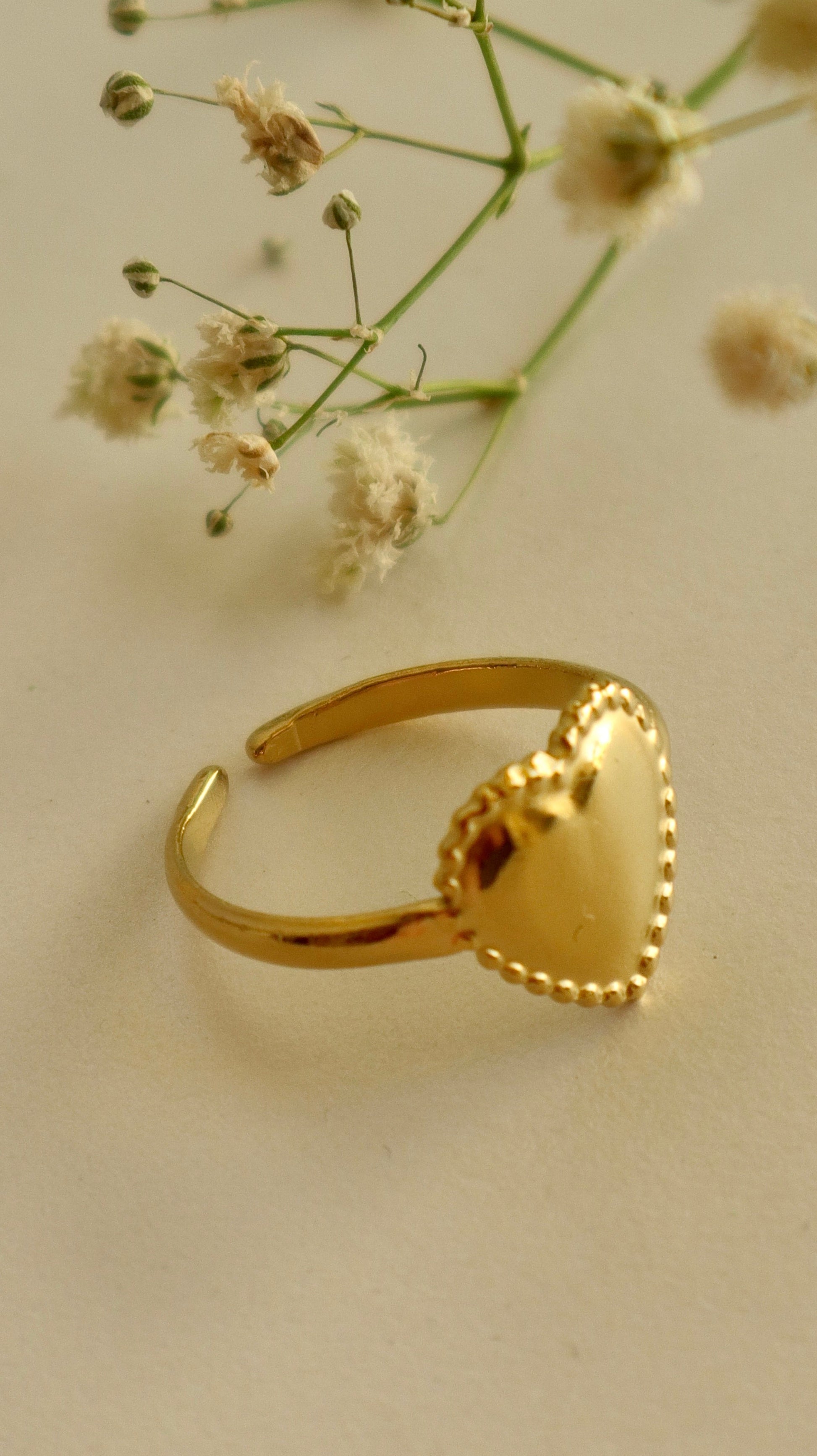 A gold-colored heart-shaped ring placed on a beige surface with small white flowers in the background.