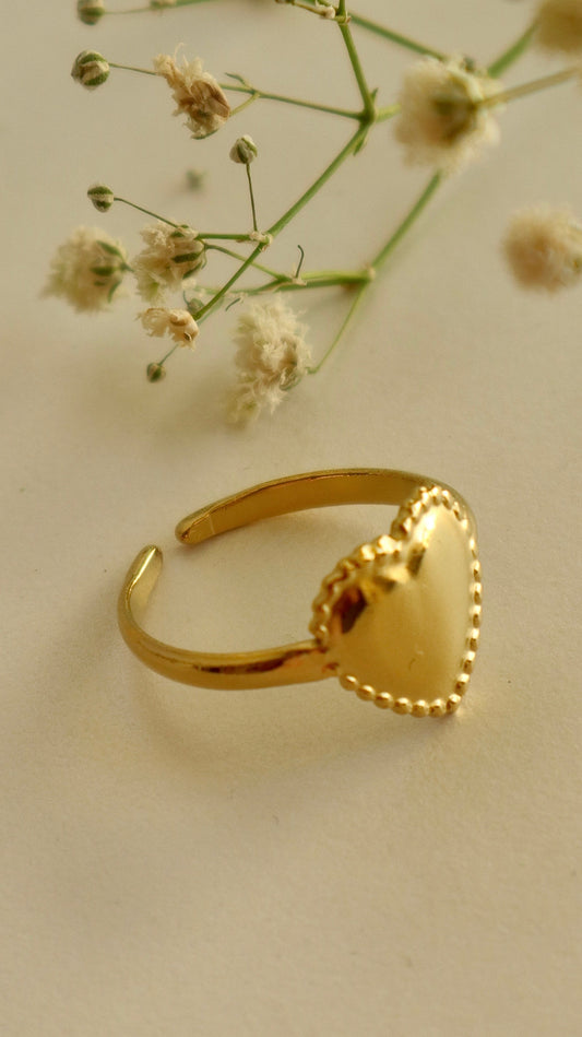 A gold-colored heart-shaped ring placed on a beige surface with small white flowers in the background.