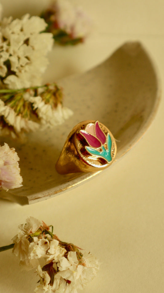 A gold ring with a bold tulip signet design, placed on a white surface surrounded by delicate white flowers.
