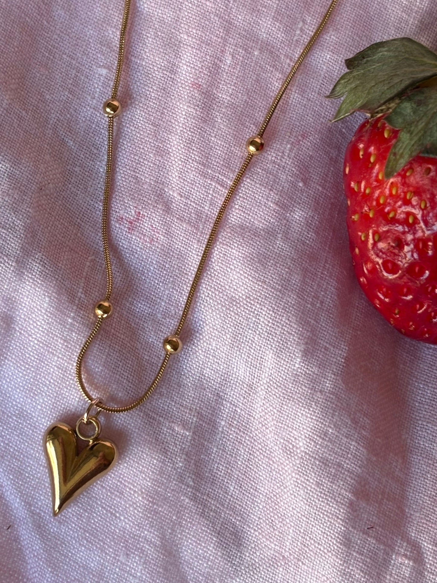 Gold beads necklace with a heart pendant on a pink fabric background with strawberries.