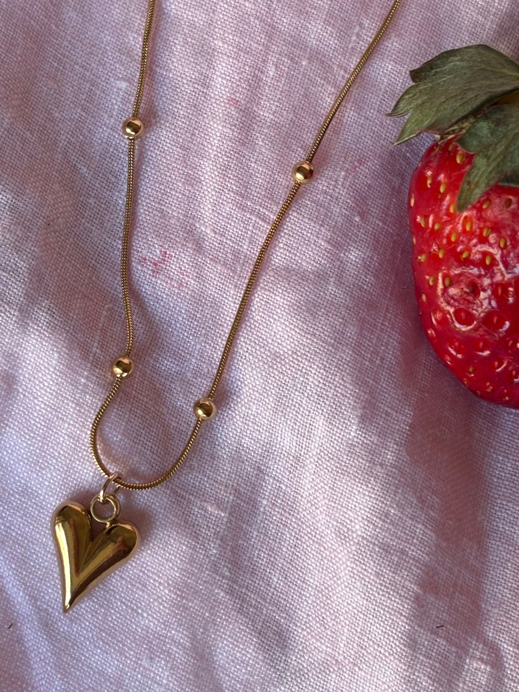 Gold beads necklace with a heart pendant on a pink fabric background with strawberries.