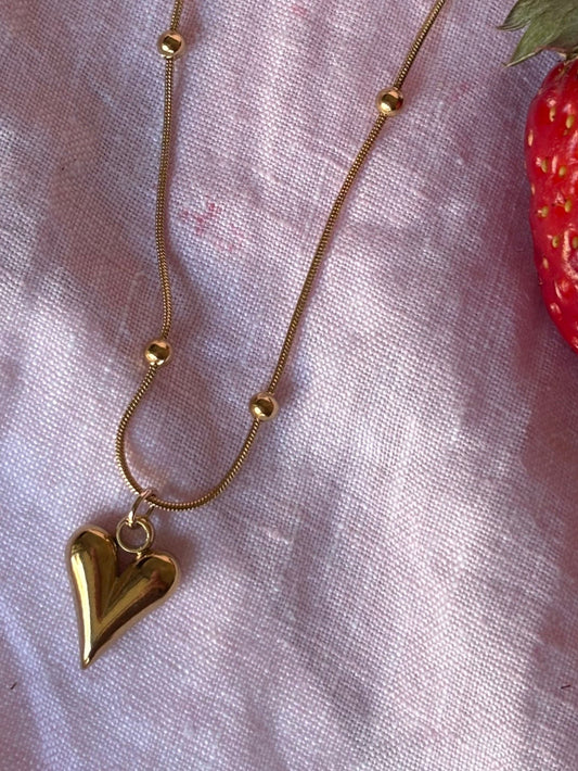 Gold beads necklace with a heart pendant on a pink fabric background with strawberries.