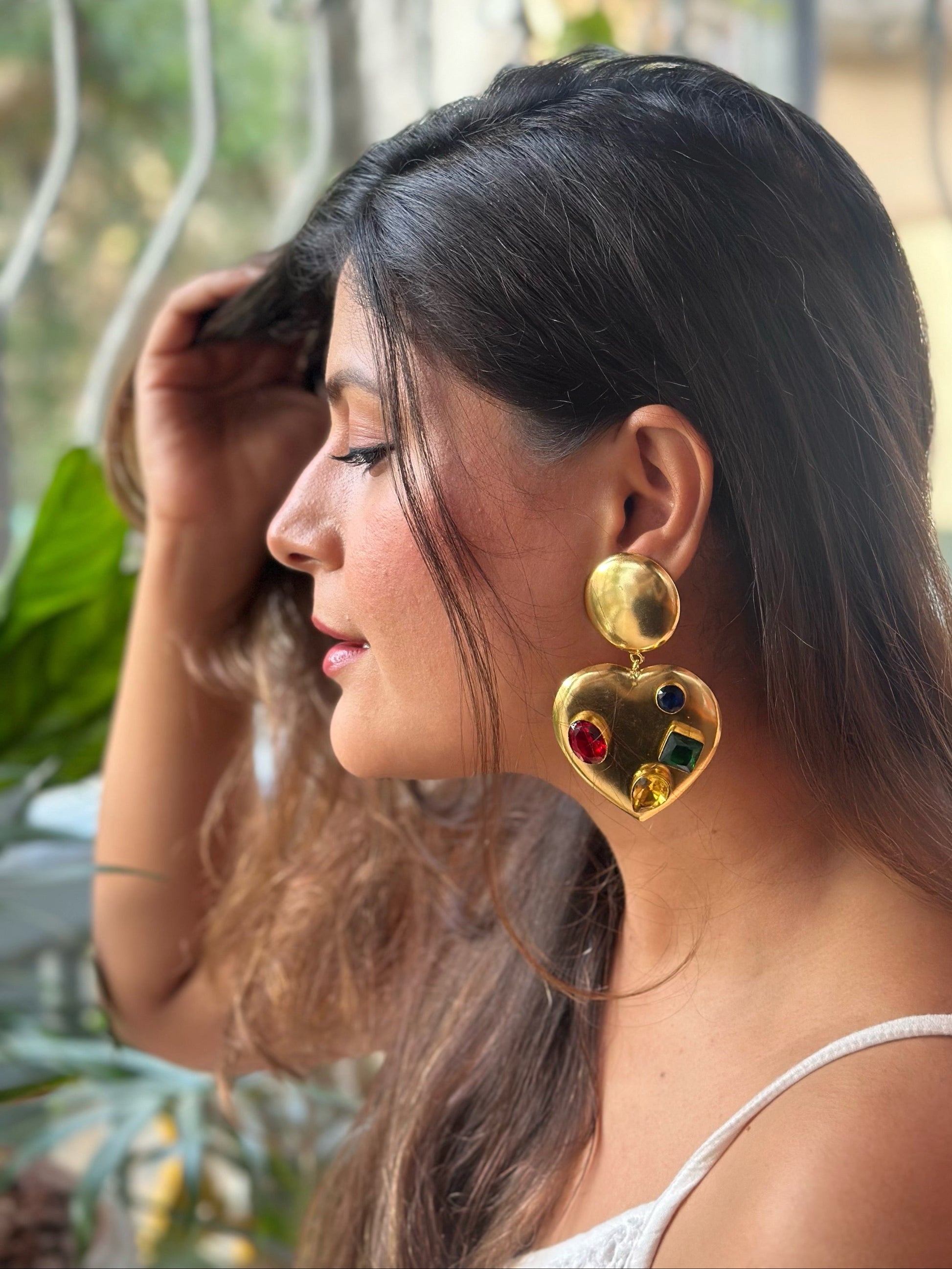Woman wearing gold heart-shaped earrings with red stones, outdoors.