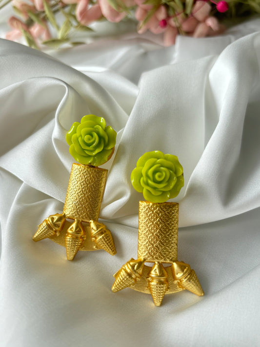 A pair of gold-colored brass earrings with a neon flower motif, displayed on a white fabric surface.