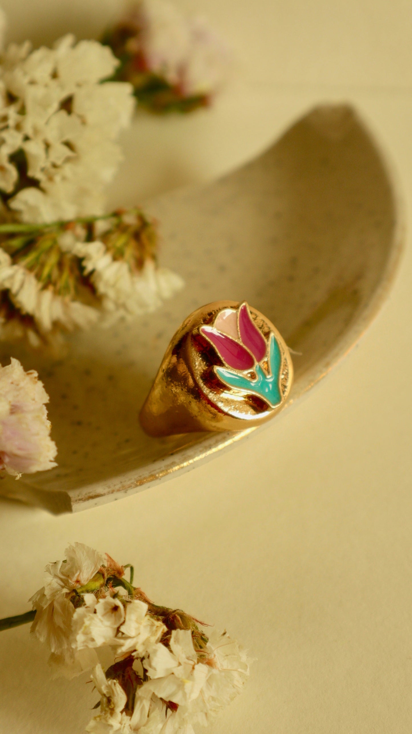 A gold ring with a bold tulip signet design, placed on a white surface surrounded by delicate white flowers.